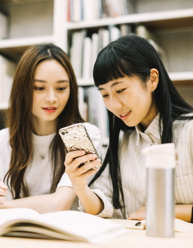 写真：図書館で一緒にスマートフォンを見る2人のアジア人女性学生