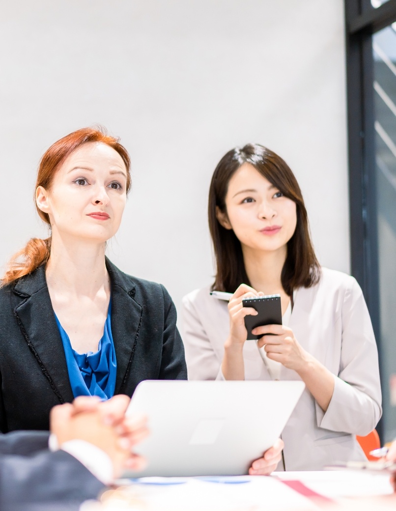 写真：オフィスでミーティングをする2人の女性ビジネスプロフェッショナル。1人は白人で1人はアジア人