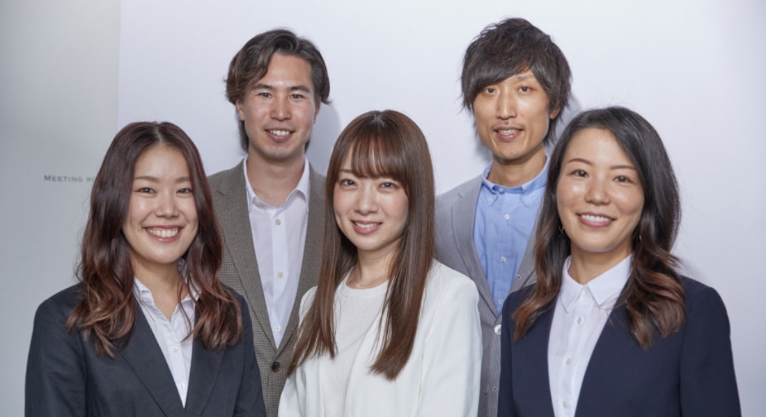 写真：ビジネススーツを着た5人のグループ。白い背景の前でそれぞれが微笑み、プロフェッショナルな雰囲気を醸し出している。