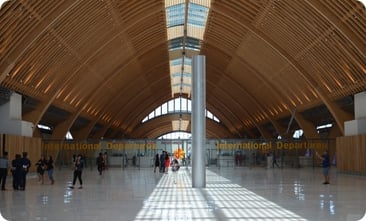 写真：大きな木造アーチの天井が特徴的な、国際線出発ゲートの広々とした空港ロビー。自然光が差し込み、明るく洗練された空間の中を歩く人々の姿が見られる。