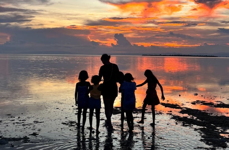 写真：夕焼けに染まる海辺で、シルエットになった大人と子供たちが手をつなぎながら波打ち際を歩く光景。オレンジ色の空と水面が反射して幻想的な雰囲気を作り出している。